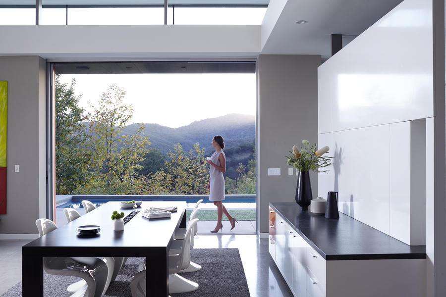 A woman holding a cup walks through a modern, open-concept home with large sliding doors that reveal a scenic mountain view and a poolside patio.